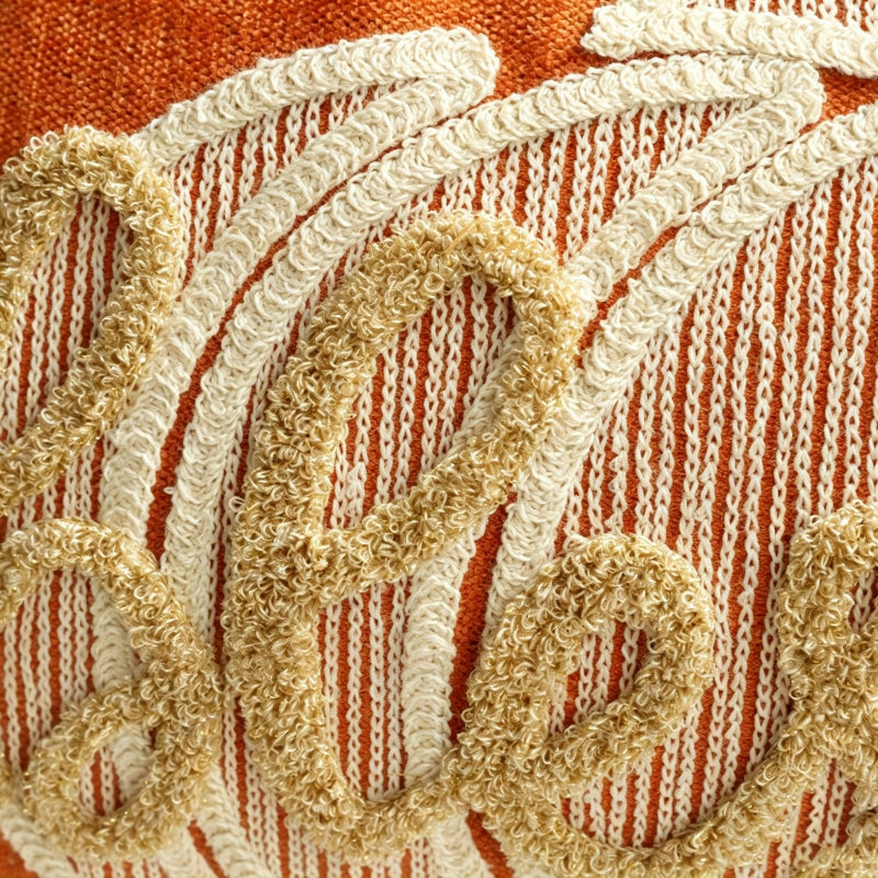 Close-up of a woven textile with a rust orange and cream striped background and large raised beige embroidered script lettering.
