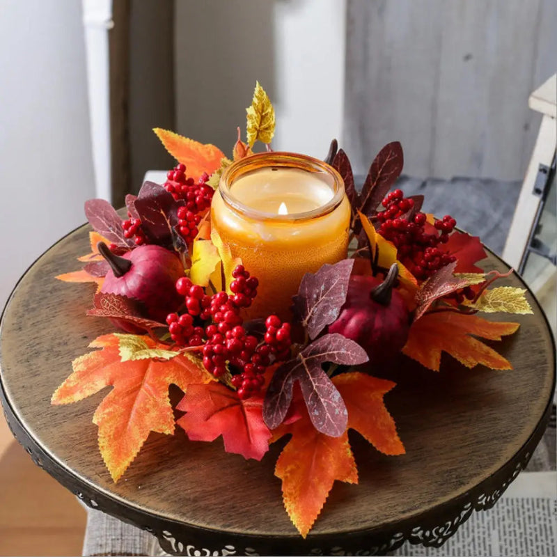 An autumn-themed candle centerpiece on a round wooden table, featuring a lit amber glass jar candle surrounded by orange and red maple leaves, red berry clusters, faux pumpkins, and a small gold leaf accent.