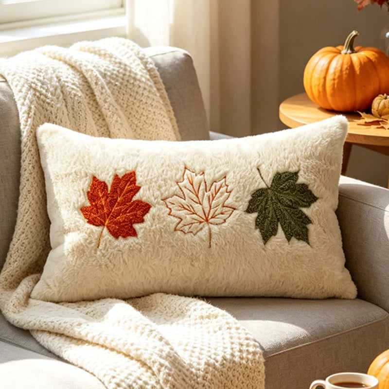 A cream-colored textured throw pillow with embroidered autumn maple leaves in red, orange, and green, styled on a gray armchair with a chunky knit cream blanket and fall decor in the background.