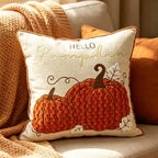 A square decorative throw pillow with a warm autumn design, featuring the text 'HELLO Pumpkin' and two textured orange pumpkins on an off-white background, styled on a beige sofa with a cozy knit blanket.