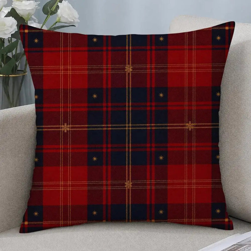 A square throw pillow with a red and navy blue tartan plaid pattern featuring small gold star details, resting on a light beige sofa next to a vase of white roses.