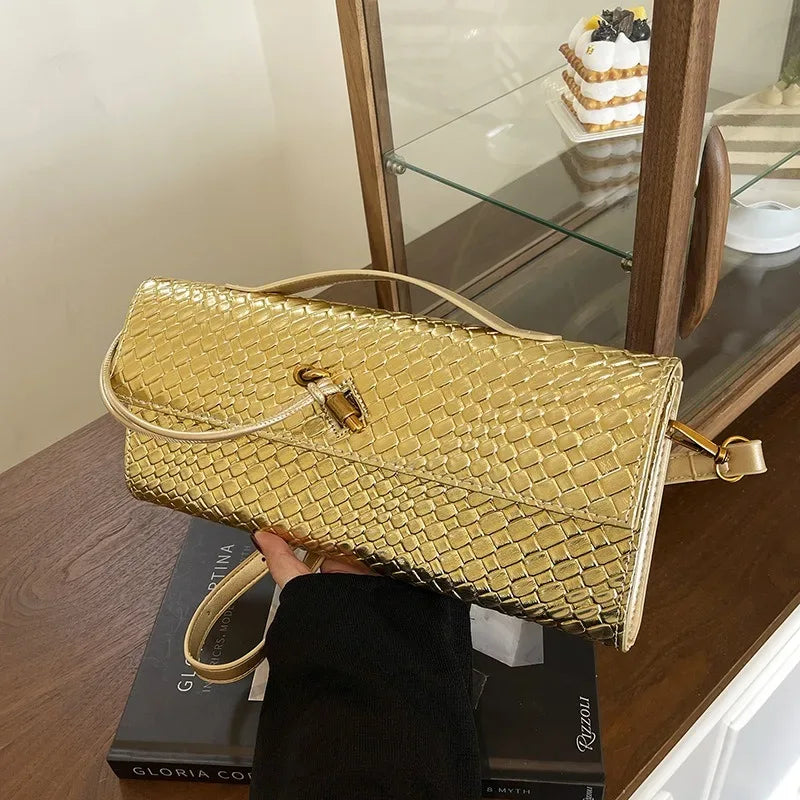 Gold woven handbag held by a person on a wooden surface with books and a glass cabinet in the background.