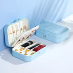 A light blue jewelry organizer box shown open and closed on a clean white and light blue studio background. The open box displays a white interior with rings, earrings, and a necklace holder, while the closed box shows the exterior with a snap closure.