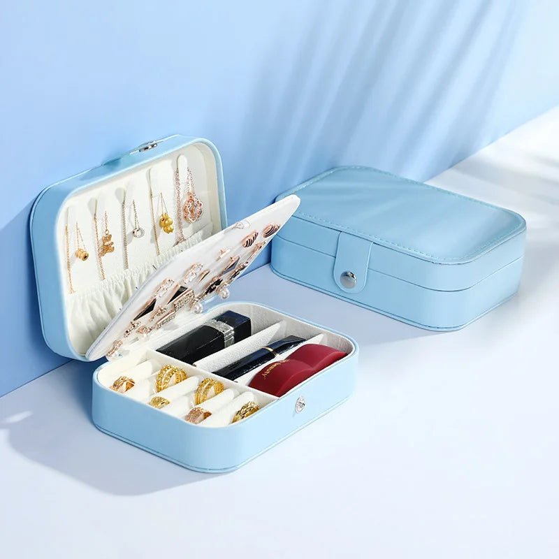 A light blue jewelry organizer box shown open and closed on a clean white and light blue studio background. The open box displays a white interior with rings, earrings, and a necklace holder, while the closed box shows the exterior with a snap closure.