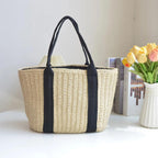 Woven bag with black straps on a white surface next to a vase of flowers and books.