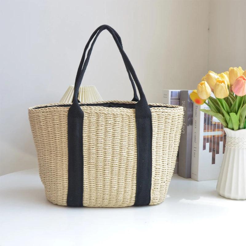 Woven bag with black straps on a white surface next to a vase of flowers and books.