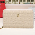 A cream-colored zip-around wallet with an embossed monogram pattern and a small gold logo at the center, displayed on a white surface with a blurred city street background.
