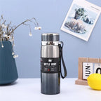 Black insulated bottle with silver lid on a white surface against a light blue wall.