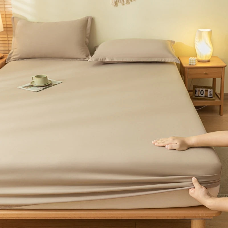 A beige fitted sheet and matching pillowcases on a bed in a warm, softly lit bedroom. Two hands are visible at the bottom edge pulling the sheet tightly, with a wooden nightstand and lamp on the right.