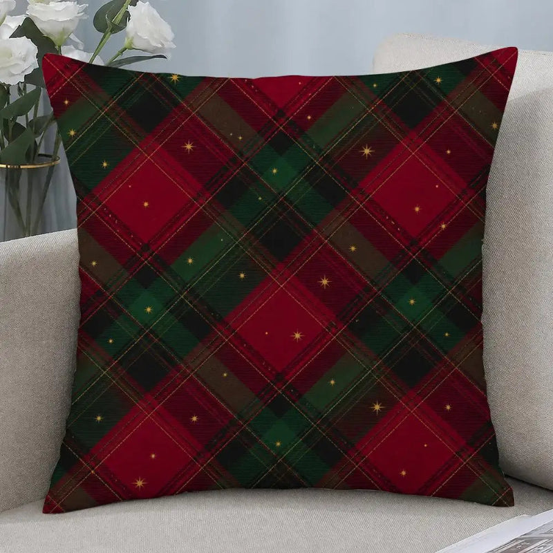 A square throw pillow with a red and green Christmas plaid pattern and small gold star accents, resting on a light beige sofa next to a vase of white roses.