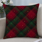 A square throw pillow with a red and green Christmas plaid pattern and small gold star accents, resting on a light beige sofa next to a vase of white roses.