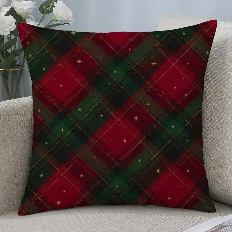 A square throw pillow with a red and green Christmas plaid pattern and small gold star accents, resting on a light beige sofa next to a vase of white roses.