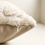Close-up of a plush cream sherpa or faux-fur cushion cover layered over a beige pillow, with a small white pull tab visible at the edge, set against a soft neutral indoor background.
