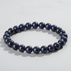 A beaded stretch bracelet made of glossy dark blue gemstone beads with tiny gold speckles, displayed on a wood slice. The text 'Self-Love' and 'Promote self-acceptance' is overlaid on the image.