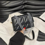 Black quilted handbag with chain strap held by a person against a black leather couch.