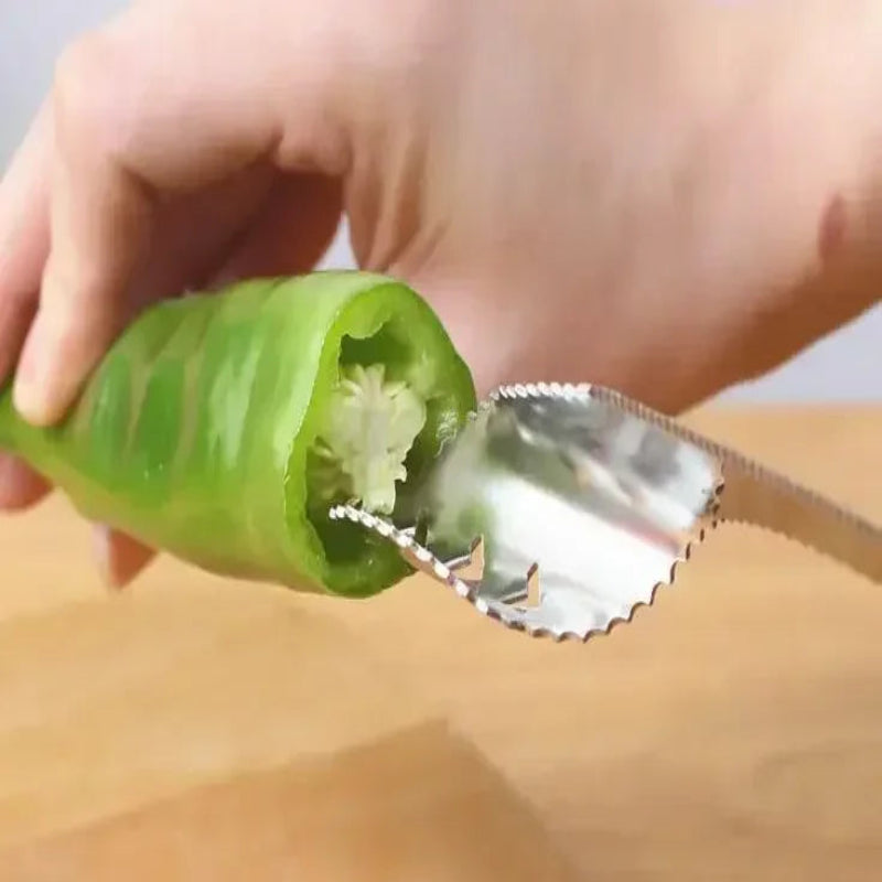 A close-up of a hand using a silver serrated metal chili pepper skinning tool to remove the inner core from a green chili pepper over a wooden cutting board.