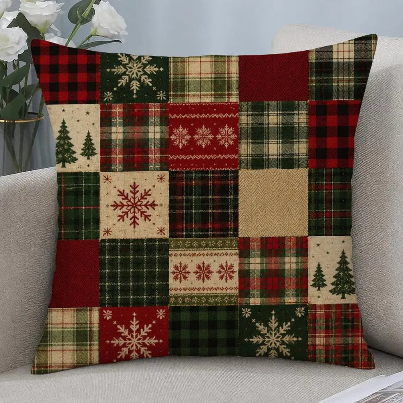 A square throw pillow with a patchwork Christmas pattern, featuring red, green, beige, and black plaid, snowflakes, pine trees, and herringbone designs, resting on a light gray sofa.