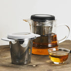 A clear glass teapot with a metal infuser and black lid sits on a wooden surface, alongside a stainless steel mesh tea infuser with a white lid and a small glass cup of amber tea.