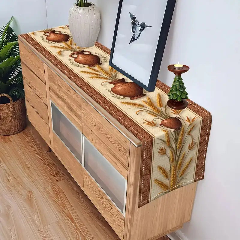 A decorative table runner with a wheat and floral pattern draped over a light wood sideboard in a room setting.