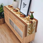 A decorative table runner with a wheat and floral pattern draped over a light wood sideboard in a room setting.