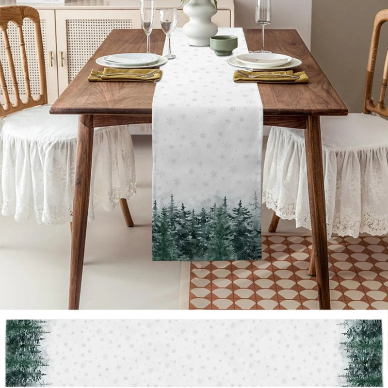 A white table runner with a green pine forest pattern at the bottom, displayed on a wooden dining table with plates, glasses, and chairs in a dining room setting.