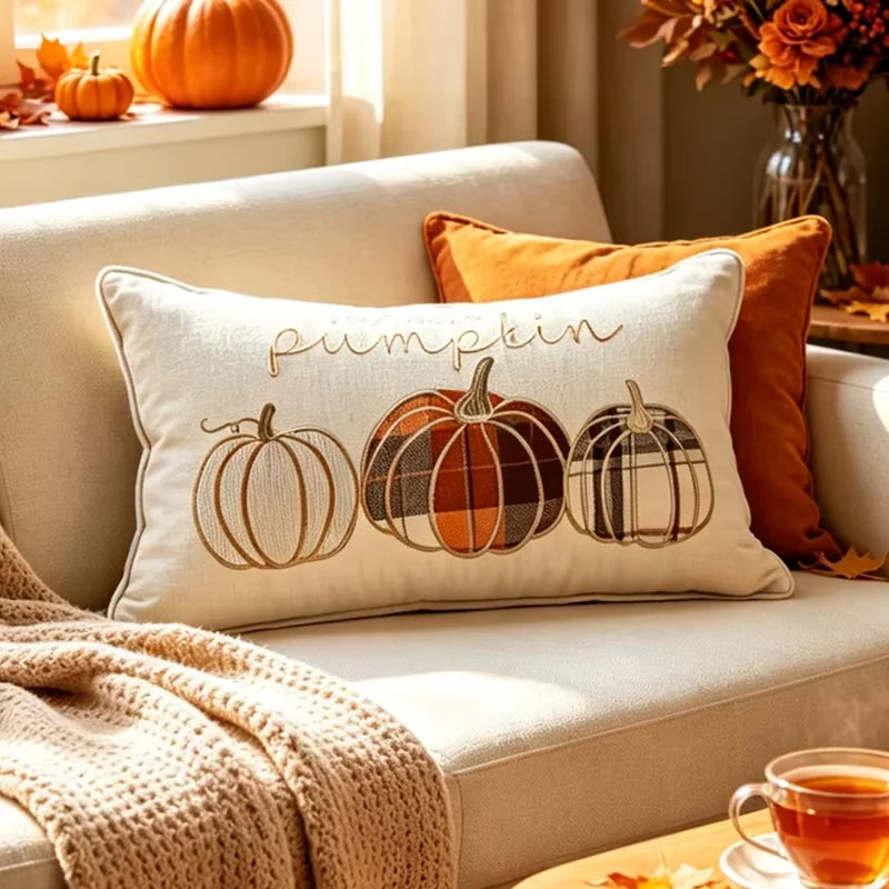 A cream rectangular throw pillow with embroidered autumn pumpkins and the word 'pumpkin' in script, styled on a beige sofa with orange and tan pillows in a warm fall living room setting.