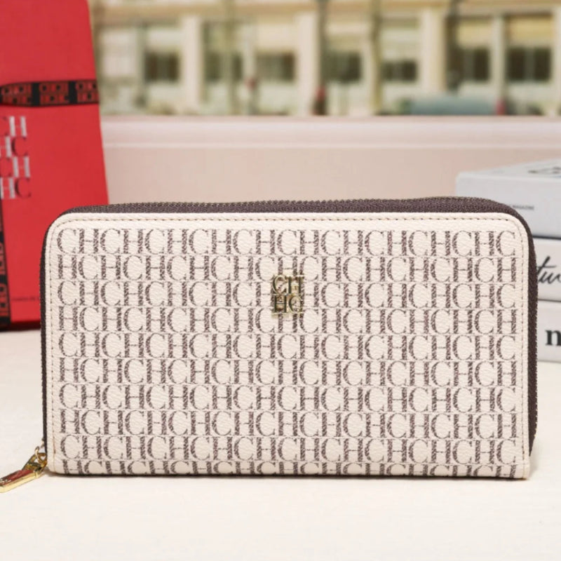 A long zip-around wallet with an allover repeating monogram pattern in beige and brown, featuring a small gold-toned logo on the front, displayed on a white surface with a blurred building facade in the background.