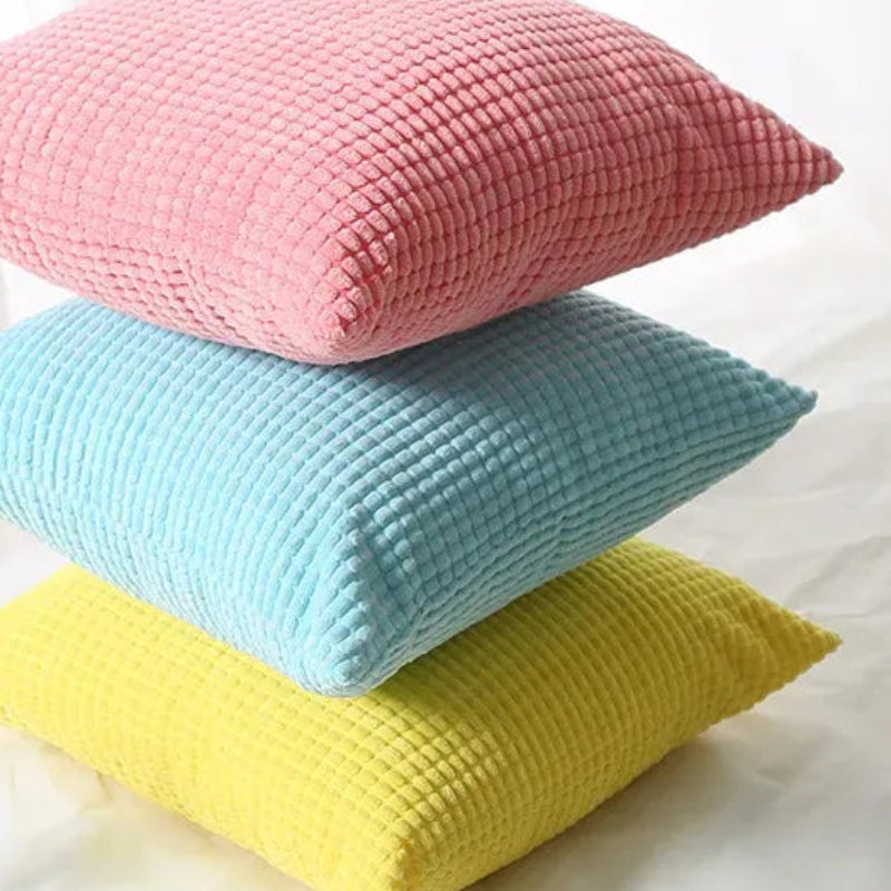Three textured pillows in pink, blue, and yellow stacked on a white surface.