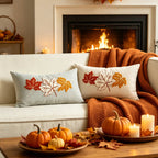 Two square throw pillows with embroidered autumn leaves in red, orange, and white, styled on a cream sofa with an orange knit blanket in front of a cozy fireplace. A wooden coffee table in the foreground holds small pumpkins, fall leaves, and lit candles.