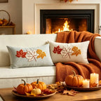 Two square throw pillows with embroidered autumn leaves in red, orange, and white, styled on a cream sofa with an orange knit blanket in front of a cozy fireplace. A wooden coffee table in the foreground holds small pumpkins, fall leaves, and lit candles.