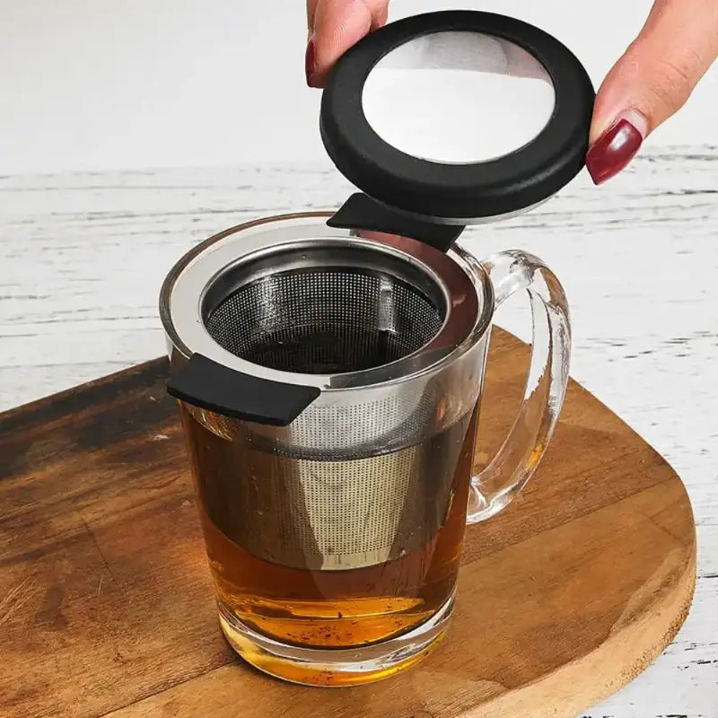 A clear glass mug with a stainless steel mesh tea infuser and black lid, shown on a wooden board with tea inside. A hand is lifting the lid, which has a clear viewing window.