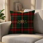 A square throw pillow with a green and red tartan plaid pattern and a central gold snowflake wreath design, resting on a light beige fabric sofa in a softly lit living room.