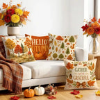 Four decorative autumn throw pillows arranged on a white sofa in a styled living room. The pillows feature fall-themed prints including trees, pumpkins, deer, leaves, and phrases like 'HELLO AUTUMN' and 'GATHER and be GRATEFUL.