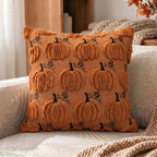 A square throw pillow in a warm rust-orange color, featuring a 3D textured pumpkin pattern with black stems and leaves, resting on a cozy beige couch with a knit blanket nearby.