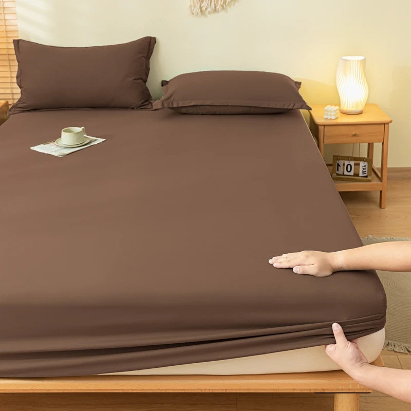 A brown fitted sheet and matching pillowcases are shown on a bed in a bedroom setting, with a person's hands pulling the sheet down over the mattress. A wooden nightstand with a lit lamp is visible in the background.