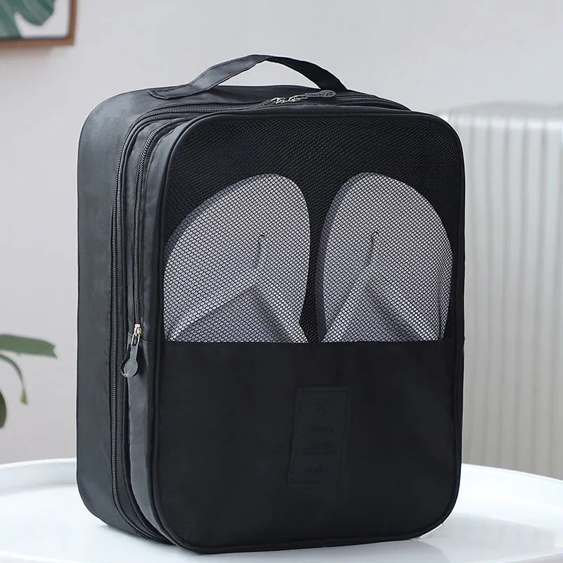 A black shoe bag with a top handle, zippered closure, and two front mesh shoe inserts, shown on a white surface against a light indoor background.