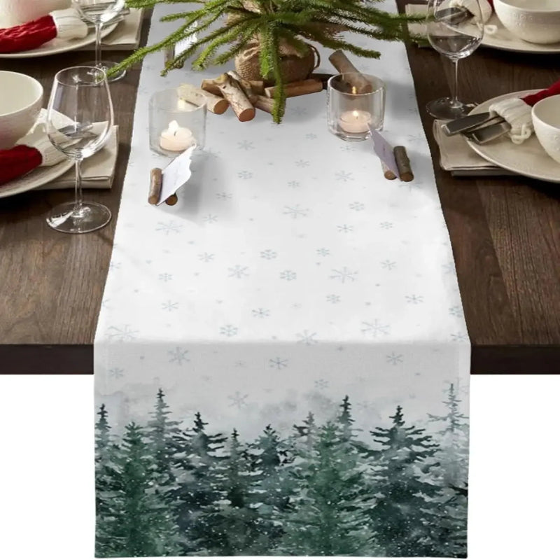 A white table runner with a snowflake pattern and a winter forest design at the bottom, displayed on a wooden dining table with place settings and holiday decor.