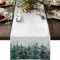 A white table runner with a snowflake pattern and a winter forest design at the bottom, displayed on a wooden dining table with place settings and holiday decor.